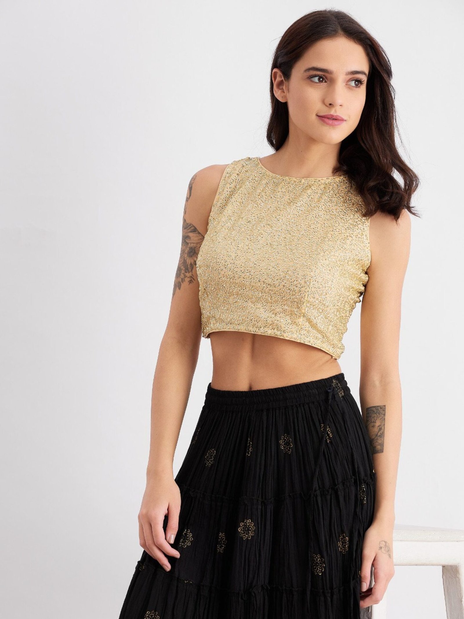Fashion photograph showing a woman in a sleeveless cropped gold top with textured fabric and a long black skirt with gold embroidered patterns, tattoos visible on her arms, posing against a plain background.