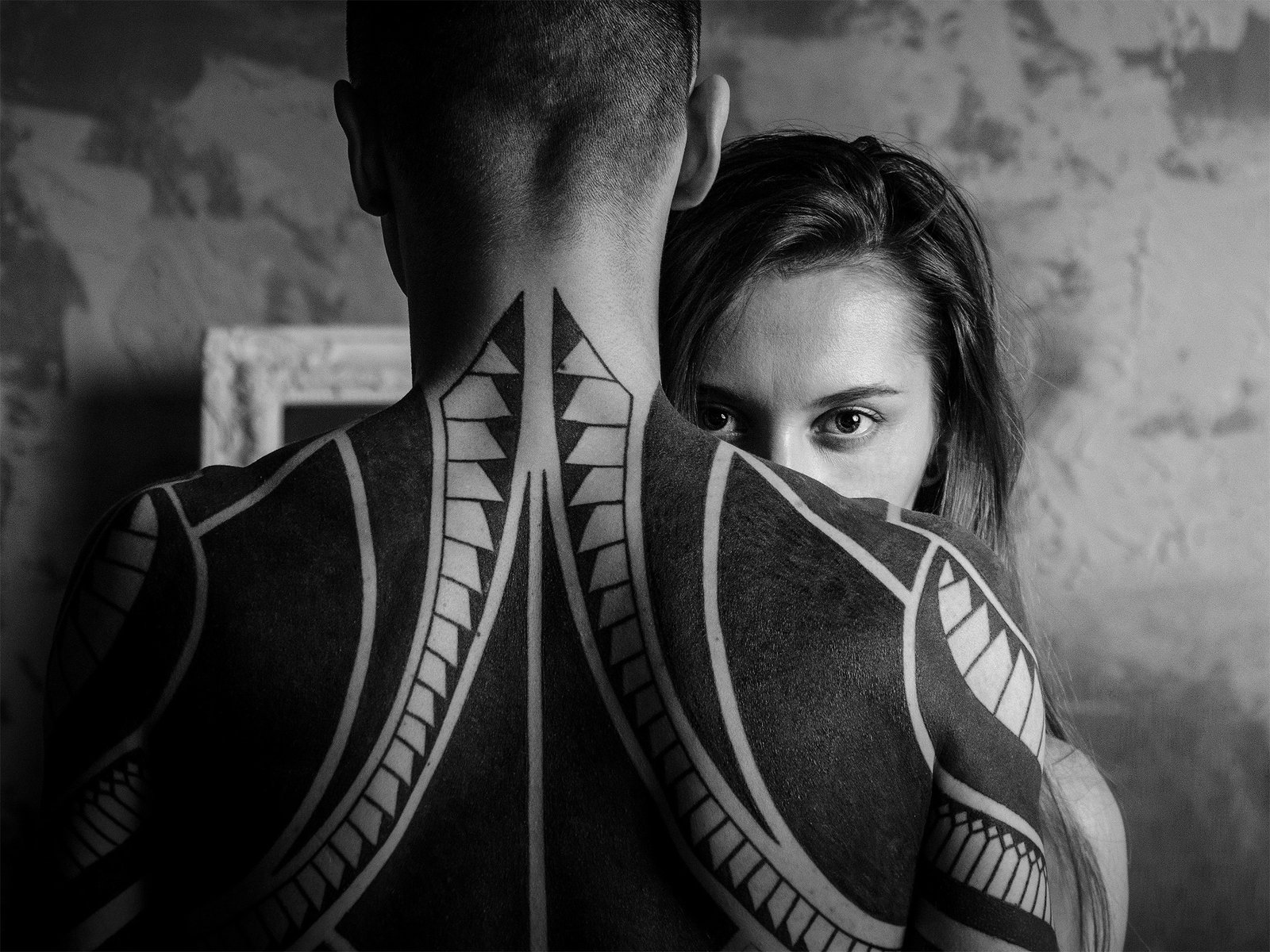 Black and white photograph showing a man with a large geometric tattoo across his upper back and shoulders, while a woman gazes intently toward the camera from behind him.