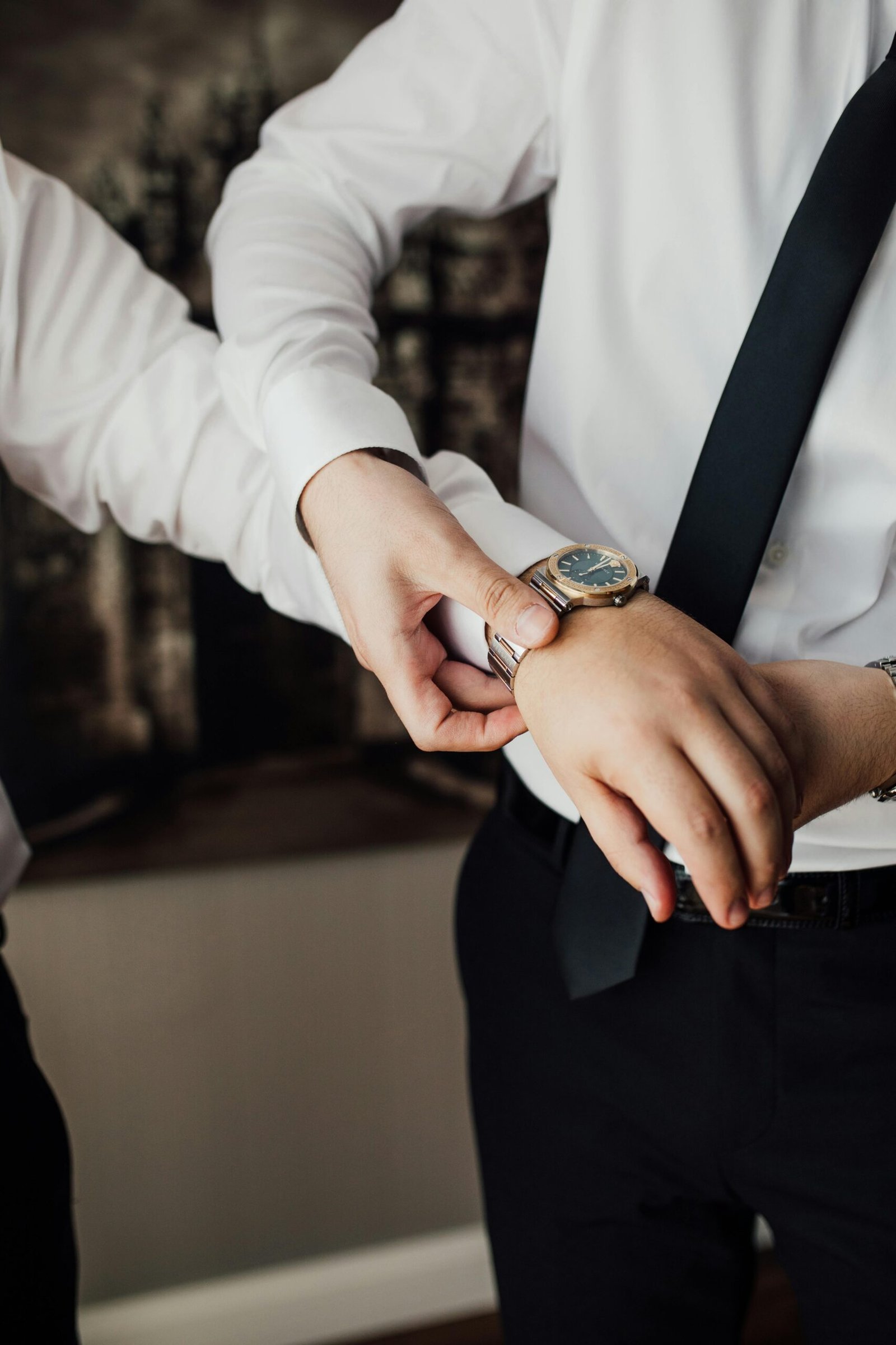 Two individuals in formal white shirts and dark trousers indoors, one fastening a metallic‑band wristwatch on the other’s wrist, capturing a moment of care and attention to detail.