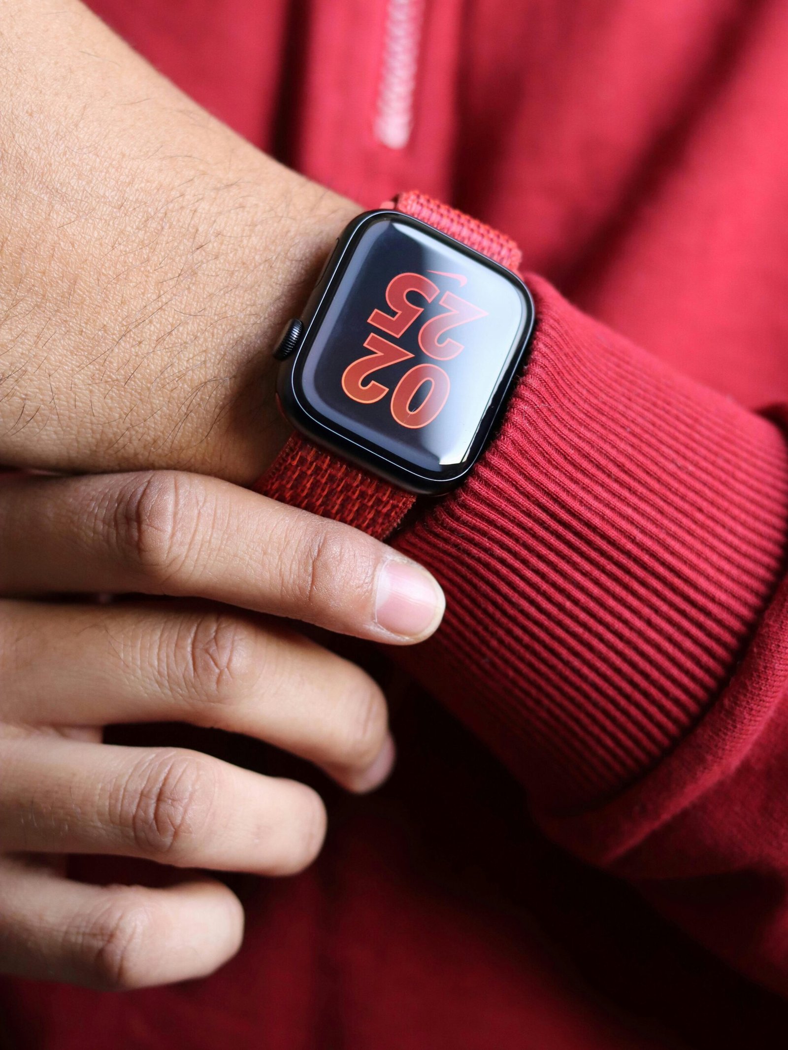 Close-up of a wrist wearing a smartwatch with a red woven band, displaying the time 02:55 in large red-orange numbers on a dark screen. The wearer’s red garment matches the watch band, highlighting a coordinated modern style.