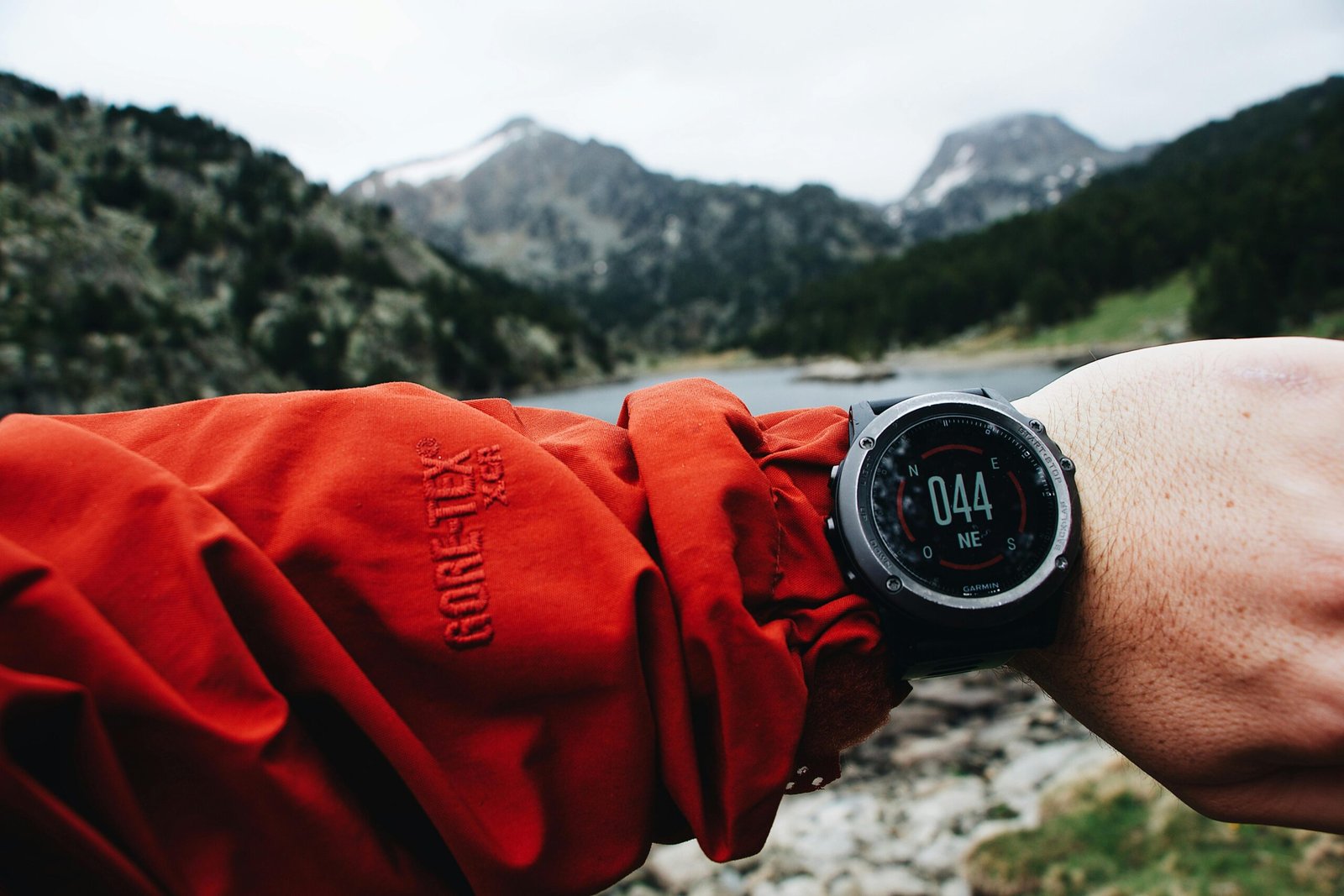 Person in a red jacket extending an arm with a Garmin watch showing compass reading 044° NE, against a backdrop of snowy mountains, lake, and forest under cloudy skies.